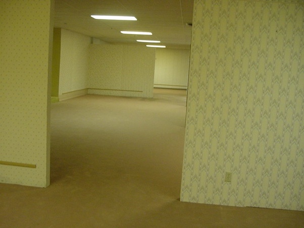 The original yellow-room Backrooms photograph, showing an empty commercial interior with fluorescent lights and patterned wallpaper.
